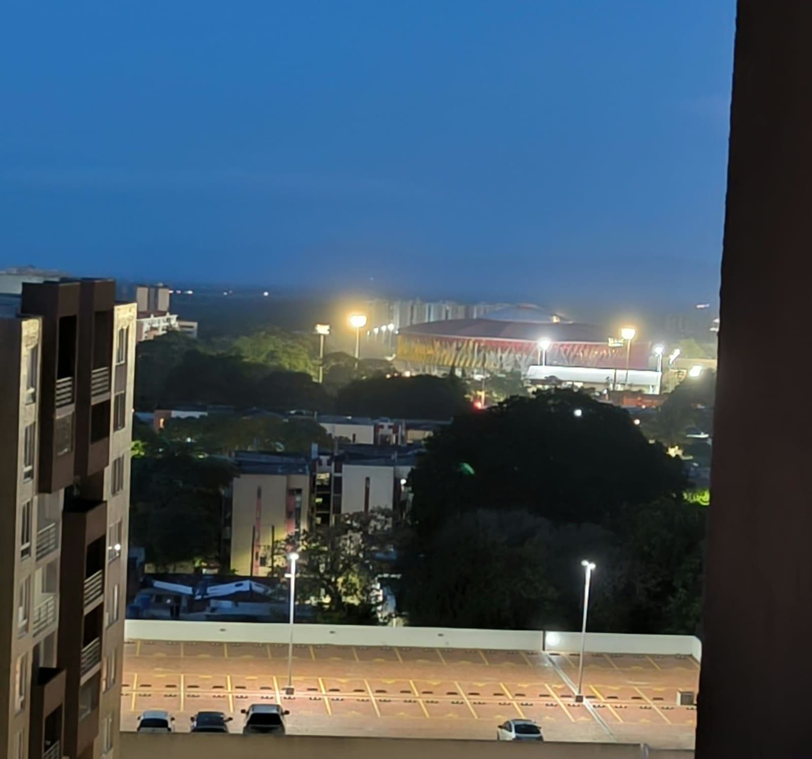 Vista nocturna desde el balcón: estadio iluminado, parqueaderos y edificios